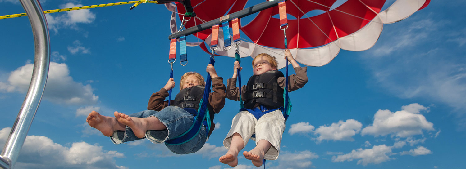 2 smiling young boys sitting tandem in parasail harness lifting into the sky