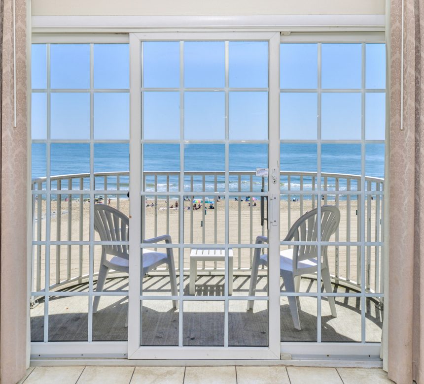 Open curtains reveal glass door and balcony with chairs overlooking beach